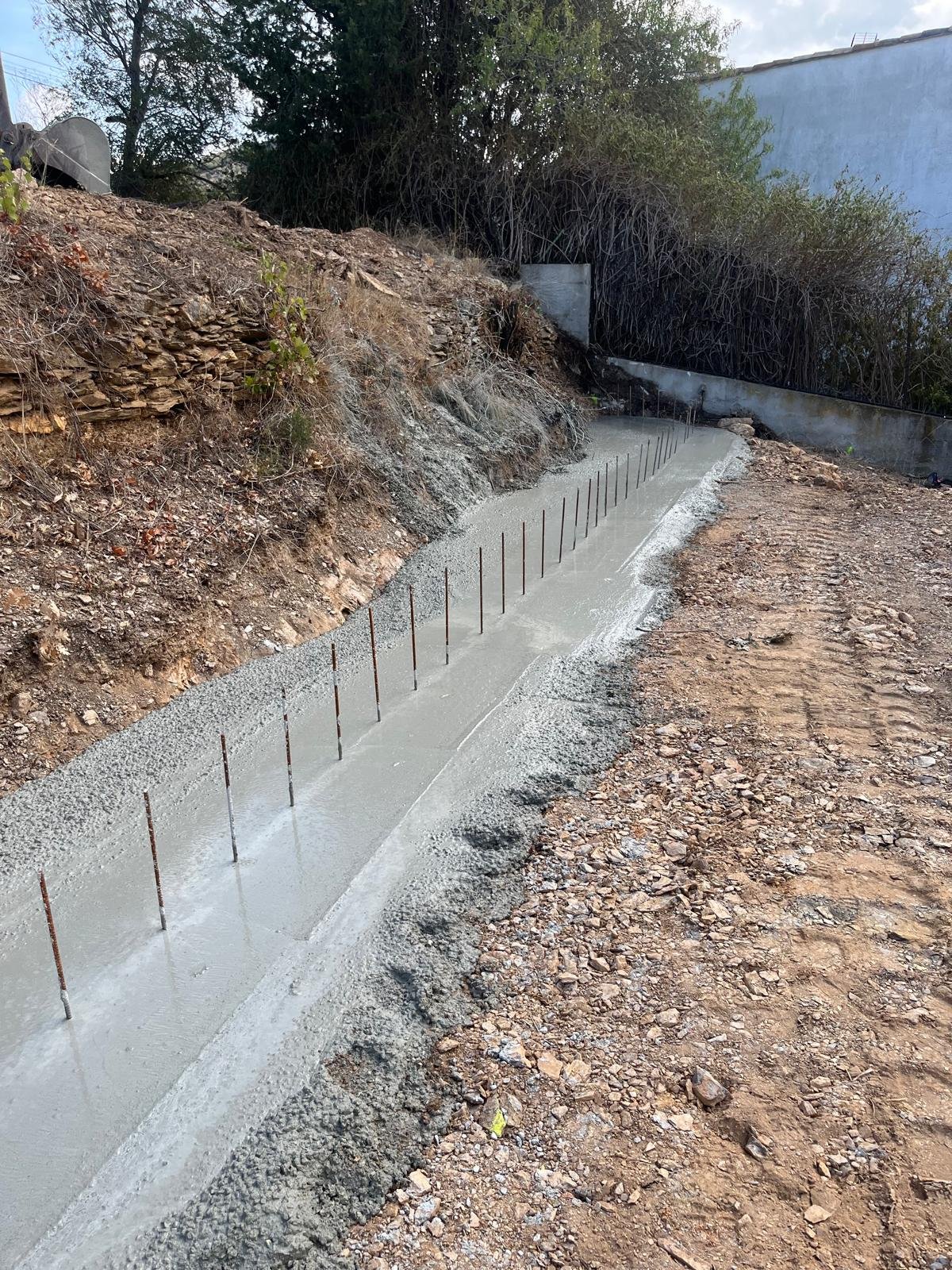 Une longue fondation en béton frais avec des barres d'armature verticales, creusée dans un terrain accidenté avec de la terre et des rochers, entourée de végétation.