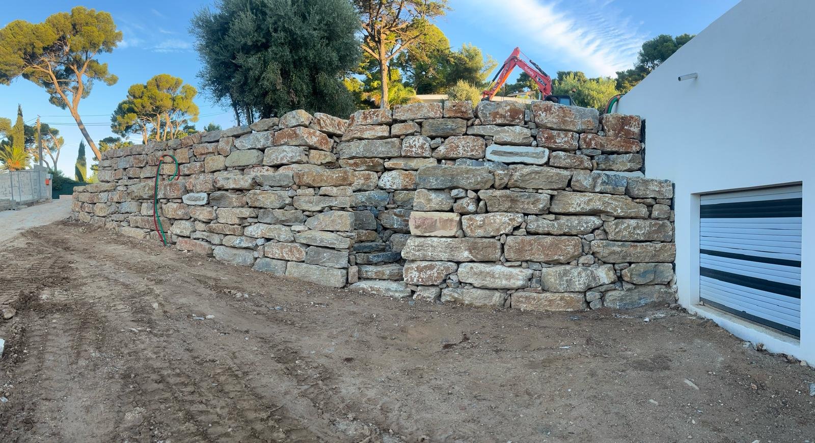 Un mur de soutènement en pierres sèches est construit devant une maison blanche et un terrain de terre, avec des arbres et un engin de chantier en arrière-plan sous un ciel bleu.