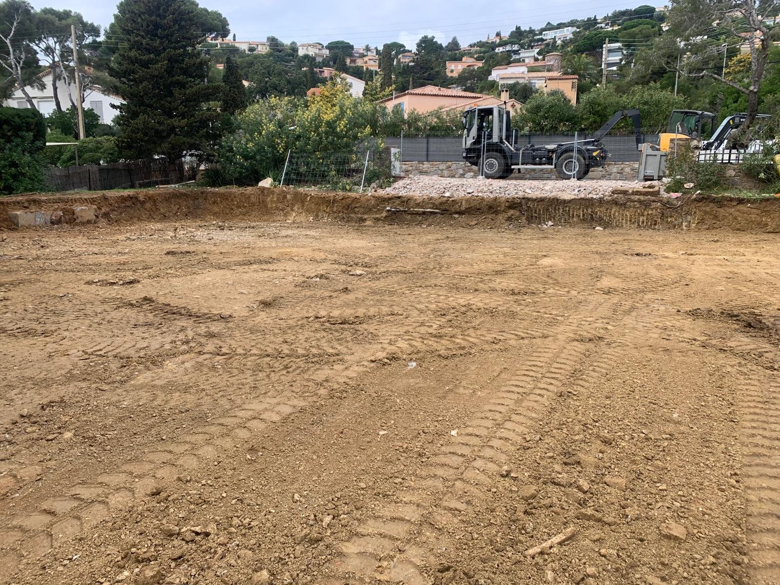 Grand terrain de chantier nivelé en terre avec des traces de pneus, un camion benne et une pelle mécanique en arrière-plan, et des maisons sur une colline boisée.